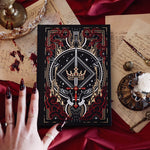 The Oath Breaker Journal sits on a red fabric background surrounded by handwritten notes, a candle and a crown. The black journal features intricate gold, red and white foiling with motifs inspired by fantasy books such as crowns, swords, goblets and snakes.