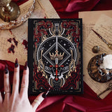 The Oath Breaker Journal sits on a red fabric background surrounded by handwritten notes, a candle and a crown. The black journal features intricate gold, red and white foiling with motifs inspired by fantasy books such as crowns, swords, goblets and snakes.