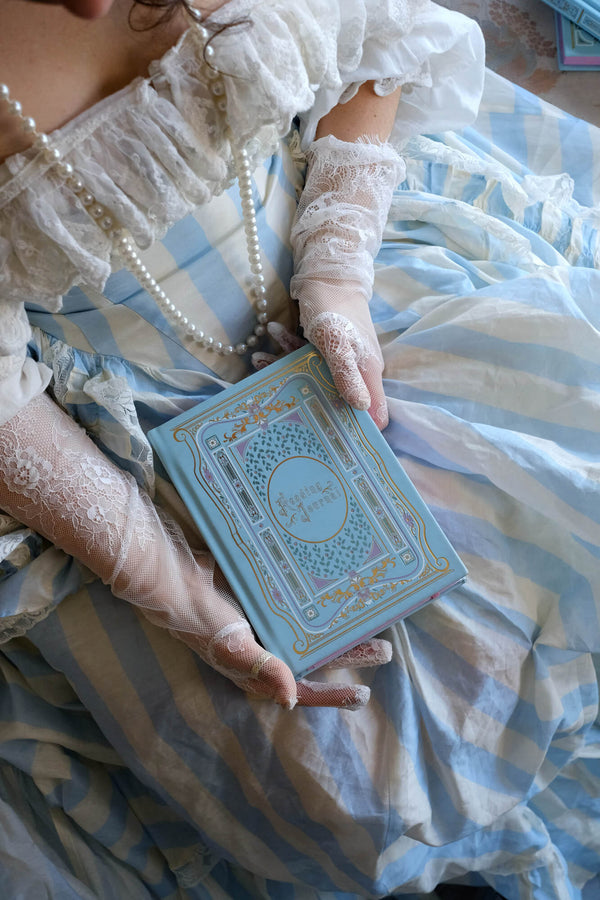 The Antoinette luxurious foiled reading journal is held by a woman in a blue striped dress with lace gloves. The aqua notebook features gold foiling and pink edge painting and is the perfect addition to the romance reader journalling aesthetic.