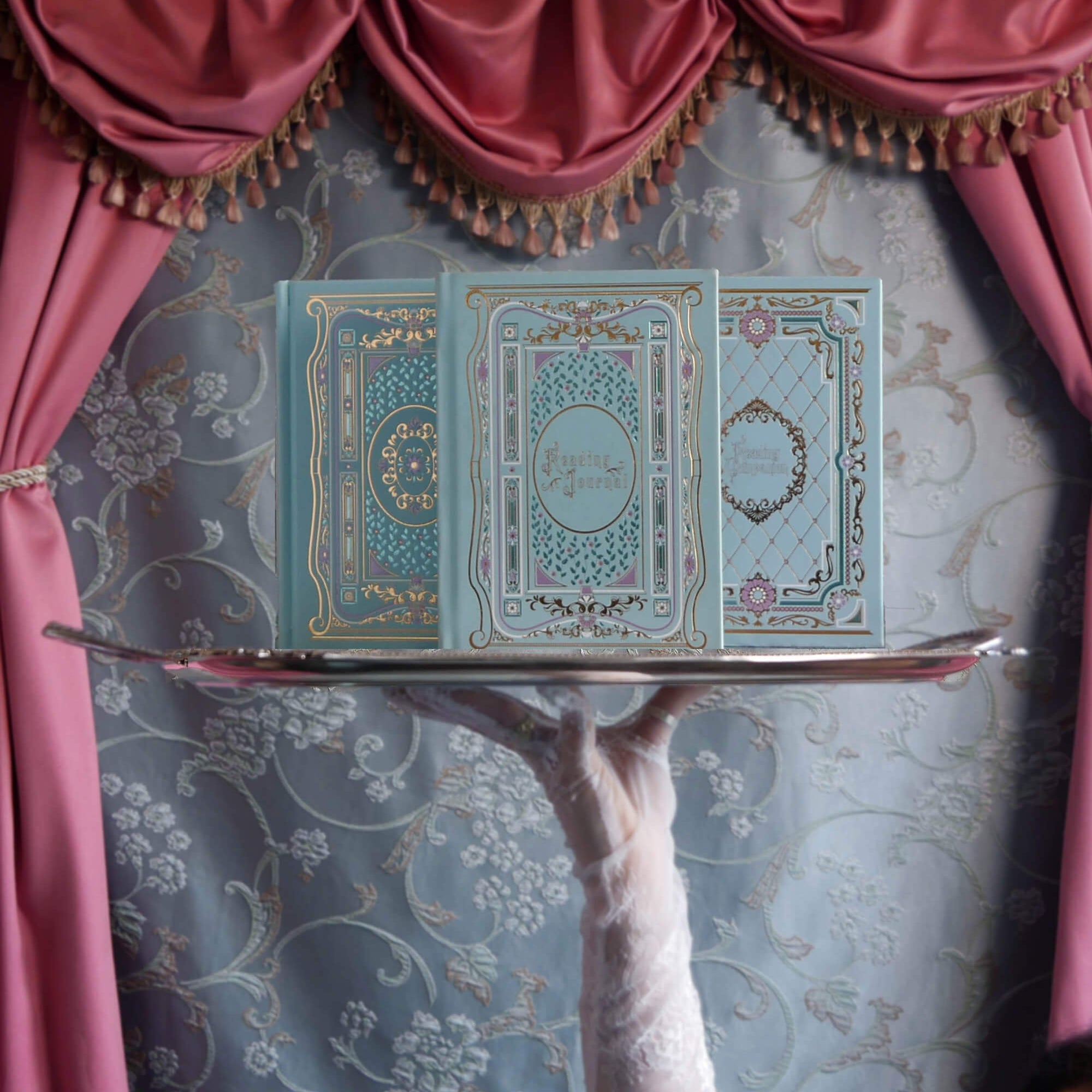 A campaign image for the Antoinette foil journal launch. In front of a blue brocade tablecloth surrounded by warm pink curtains there is a hand with lace gloves holding a silver platter. The journal, reading journal and reading companion sit atop this, their metallic foil notebook detailing catching the light.