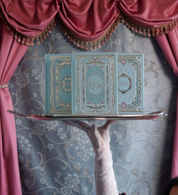 A campaign image for the Antoinette foil journal launch. In front of a blue brocade tablecloth surrounded by warm pink curtains there is a hand with lace gloves holding a silver platter. The journal, reading journal and reading companion sit atop this, their metallic foil notebook detailing catching the light.
