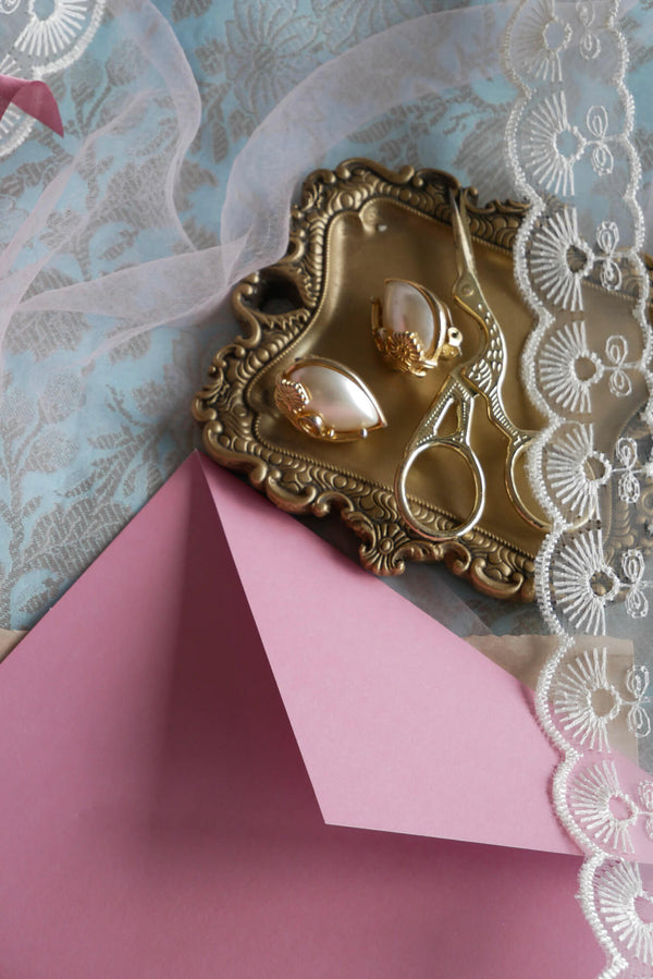 A campaign image for the Antoinette stationery launch. On a blue brocade tablecloth covered in ribbons is a gold with pearl earrings and embroidery scissors, a pink envelope to the side.
