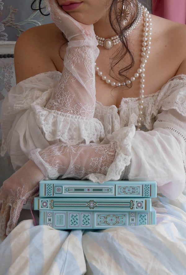 A woman leans on a stack of the Antoinette journals. She wears a white dress, peals and lace gloves and is perfectly presenting the unique foil notebooks 