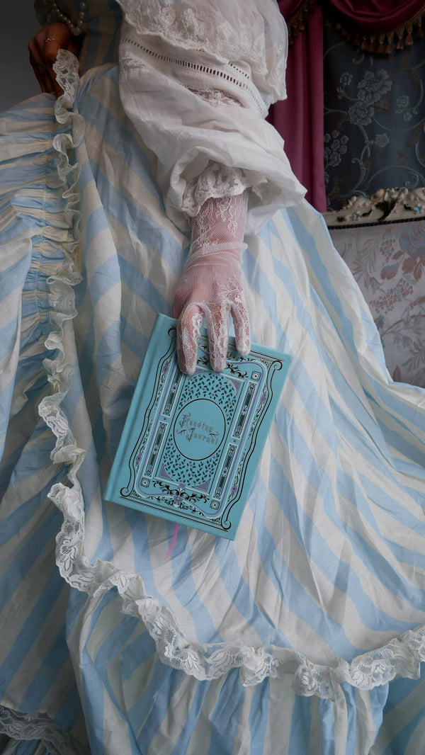 A woman in a white and light blue striped dress holds the light blue Antoinette reading journal in front of a french inspired room. The foiled notebook is inspired by a classic french journaling aesthetic.