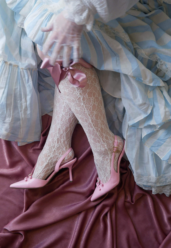 Legs wearing lace stockings and pink heels sit on a dusty pink fabric background. The woman is wearing a light blue and white striped dress as well as lace gloves for a whimsical french aesthetic.
