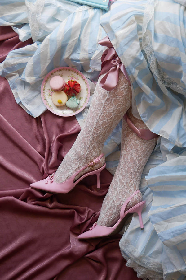 Legs wearing lace stockings and pink heels sit on a dusty pink fabric background. The woman is wearing a light blue and white striped dress and sits next to a plate with delicate French pasteries.