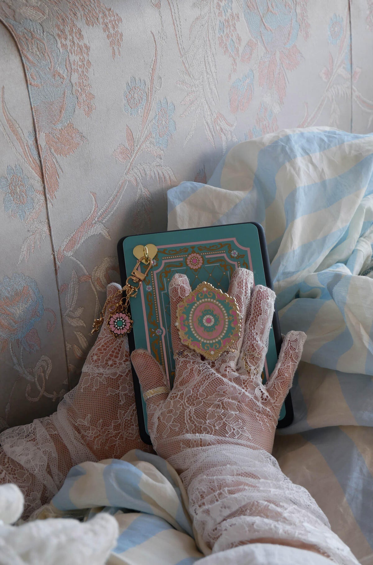 Hands wearing lace gloves hold a decorated Kindle embellished by the Antoinette eReader decor set. The clingie (think a classical book pop socket), sticker insert and charmlet create a whimsical Kobo aesthetic.