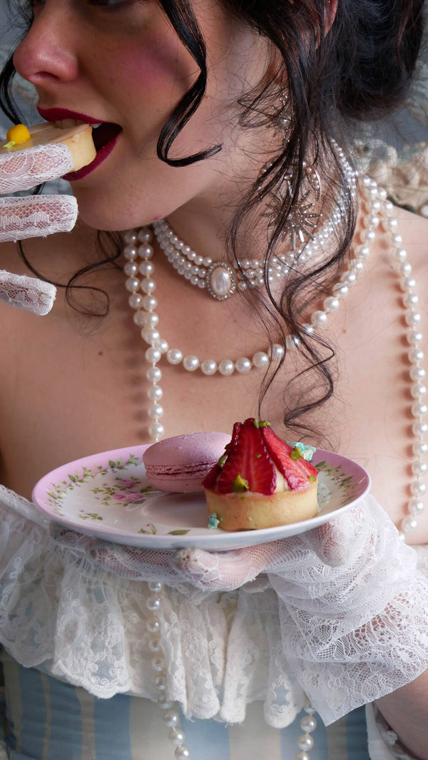 A woman wearing multiple stacks of peals and a white and blue striped dress eats french pastries from an ornate pink plate.