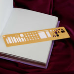The gold Metal reading journal stencil ruler by The Quirky Cup Collective sits on an open reading journal and is engraved with journaling symbols such as ratings, genre and a bookshelf on a deep pink red fabric background.