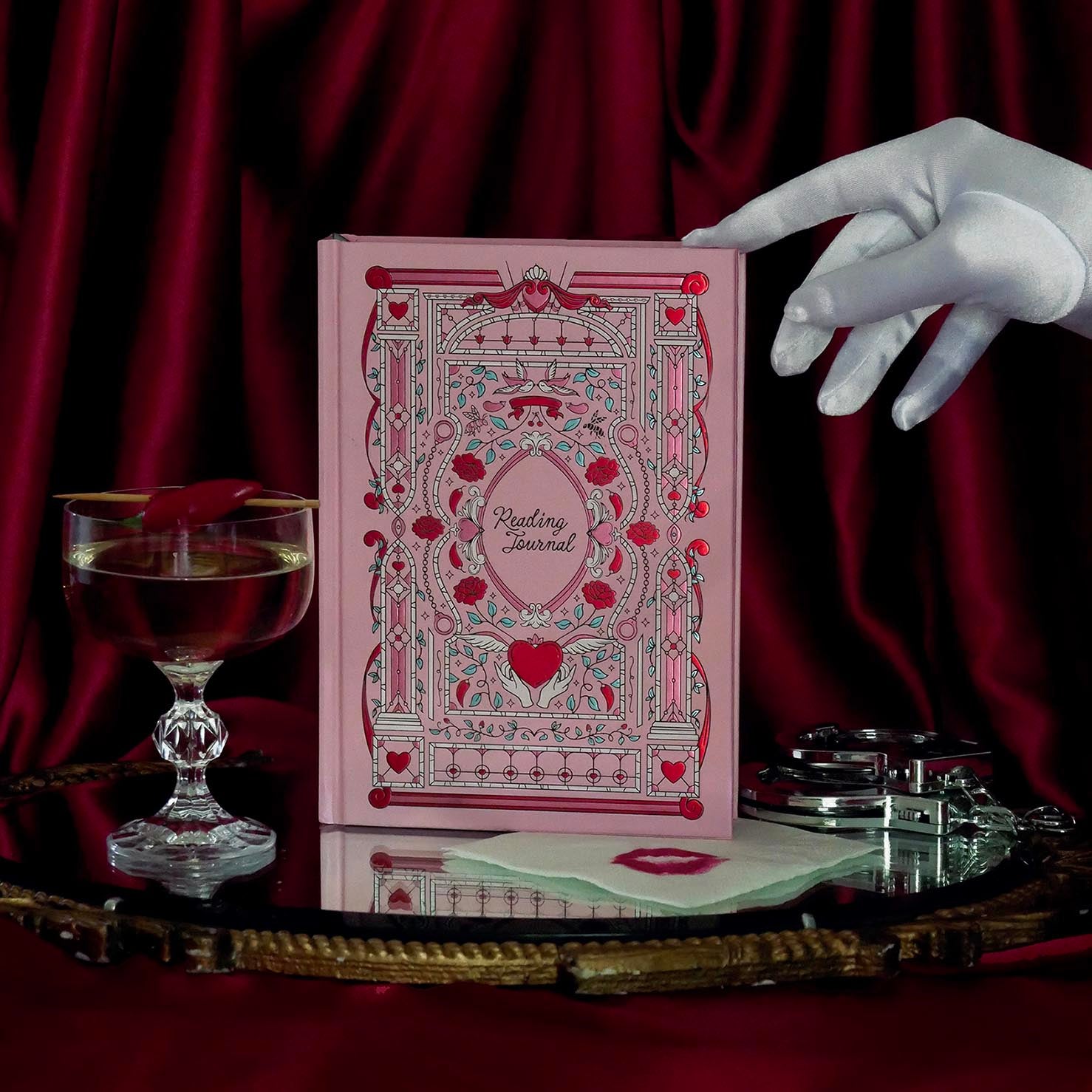 The pastel pink Sugar & Spice Reading Journal features intricate foiled details in shades of red, green, pink and white. The cover design features rose, heart, bird, handcuff, bee and flower motifs. The romance book aesthetic journal sits on a red fabric and pearls in the background.