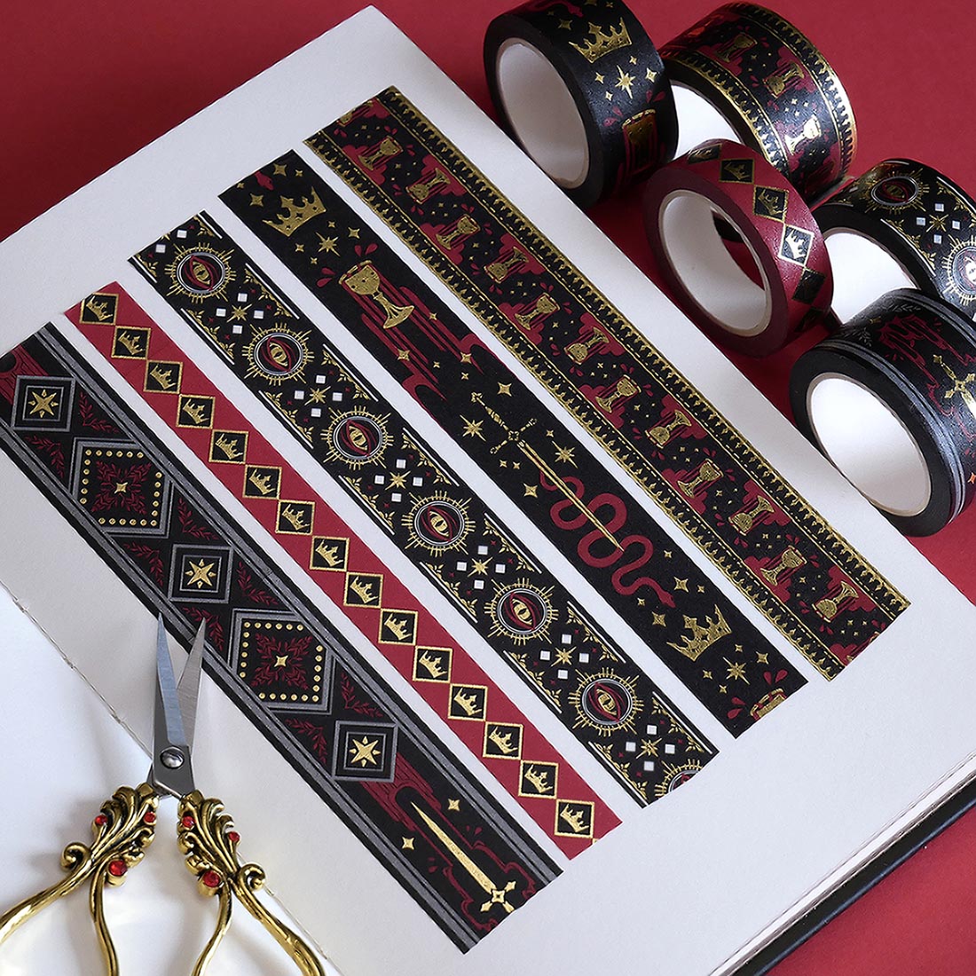 The full set of Serpents and Swords washi tapes, a romantasy book inspired collection by The Quirky Cup Collective, are show on a white journal on a red table. The five washi tapes are in shades of red, black and white with intricate gold foiling. The designs feature motifs of snakes, crowns, swords, goblets , stars and serpents and perfectly match the fantasy book aesthetic.