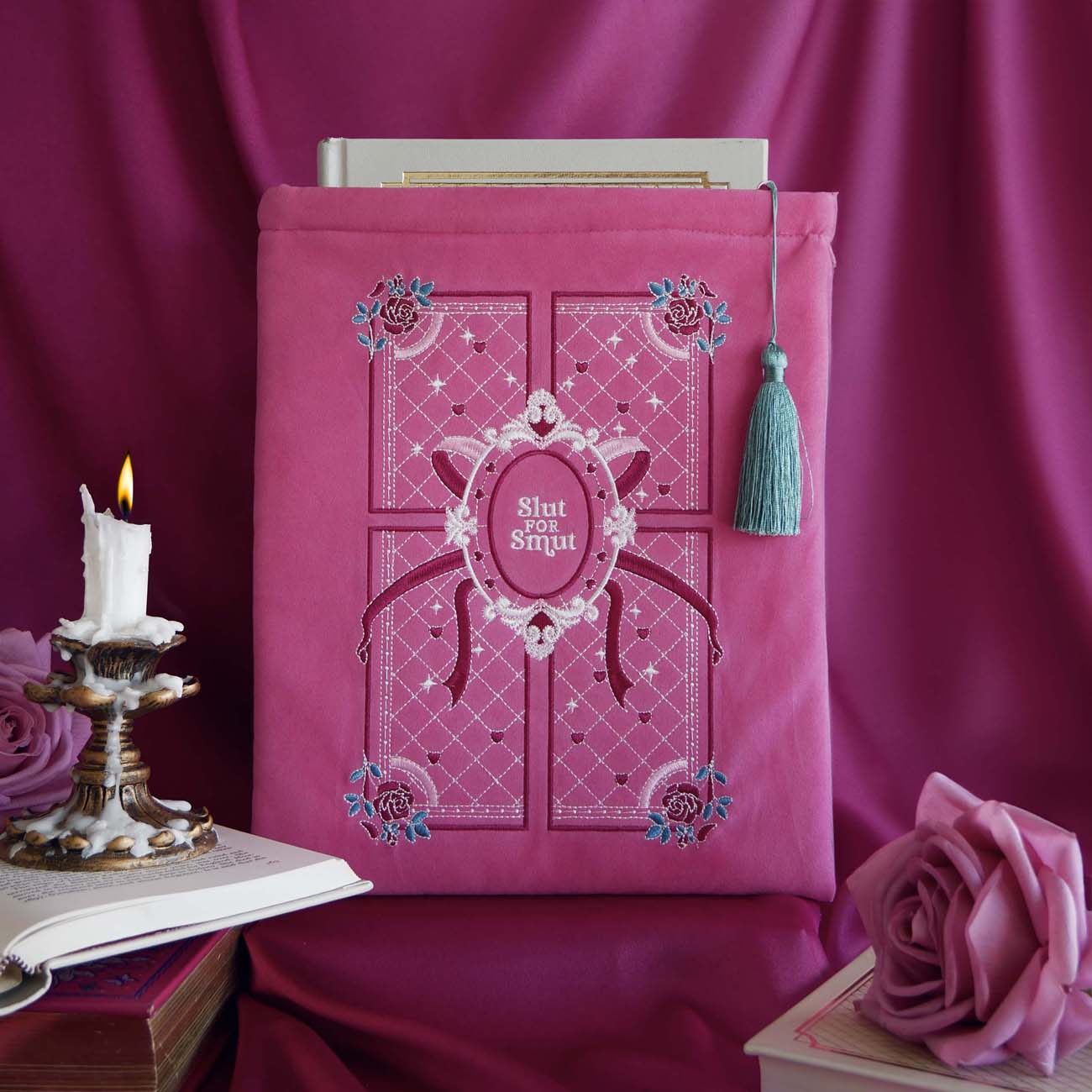 pink velvet book sleeve with pink, white and teal embroidery. Sleeve sits in front of a background of draped pink satin. Sleeve has a teal tassel and a quilted inspired design with stars, hearts, roses and bows. In the centre it says 'slut for smut'. Sleeve sits framed by a candle, books and a rose. 