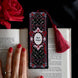 An ornate bookmark with a black, gold, and red design, featuring the text 'Slut for Smut' and a red tassel, placed on an open book with a person's hand and a red rose in the background.