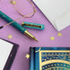 Teal 'Your are Magic' pen by The Quirky Cup Collective features gold celestial accents with the feature text engrained on the pen clip. The pen sits on a lilac background surrounded by stars, a journal and glasses.