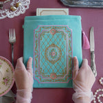 Detail of the intricate embroidery on the front of the Antoinette aqua velvet zippered Book sleeve featuring gold, pink and cream embroidery detail. The Victorian inspired design is perfect for a classic literature eReader aesthetic, shown here on a pink dining table alongside fine cutlery, peals and gloved hands.