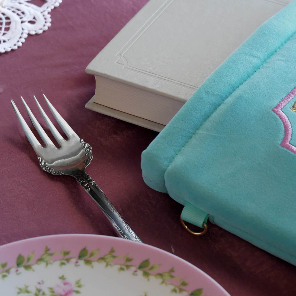 A detail of the mint green book sleeve Antoinette sits on a pink table. The aqua velvet book case features pink, cream and gold embroidery with a classic book inspired design. The 'd ring' is shown here to allow easy attaching of the wrist strap and charm elements. 