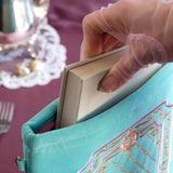 A detail of the mint green book sleeve in Antoinette. The aqua velvet book case features pink, cream and gold embroidery with a classic book inspired design. Here a hand inserts a large white book into the case, the pink satin interior showing.