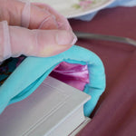 A detail of the mint green book sleeve in Antoinette. The aqua velvet book case features pink, cream and gold embroidery with a classic book inspired design. Here a hand inserts a large white book into the case, the pink satin interior showing.