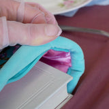A detail of the mint green book sleeve in Antoinette. The aqua velvet book case features pink, cream and gold embroidery with a classic book inspired design. Here a hand inserts a large white book into the case, the pink satin interior showing.