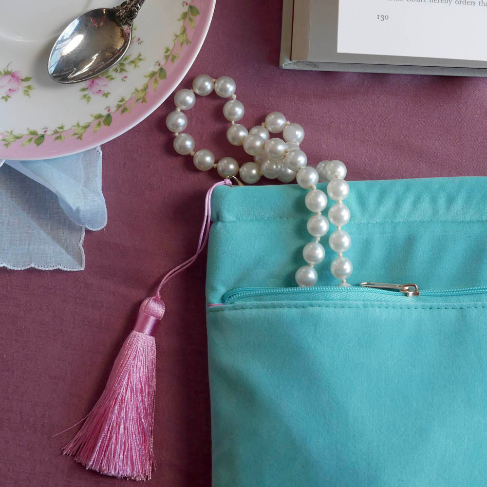 A detail of the back of the mint green book case Antoinette sits on a pink table. The aqua velvet book sleeve features pink, cream and gold embroidery with a classic book inspired design. The back pocket is shown here alongside the pastel pink tassel.