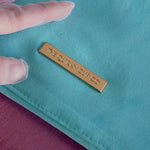 A close-up of the gold metal nameplate engraved with 'THE QUIRKY CUP CO' on the back of the aqua velvet book sleeve in Antoinette.
