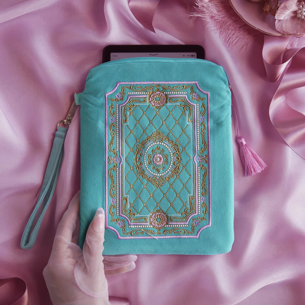The Antionette embroidered e-reader case by The Quirky Cup Collective sits on a light pink fabric background surrounded by a coquette teacup and ribbon , a hand with pastel nails holding it. The kindle case features a wrist strap and pink tassel. The mint green velvet kindle case features pink, gold and cream embroidery of a French-inspired floral frame motif. 