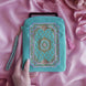 The Antionette embroidered e-reader case by The Quirky Cup Collective sits on a light pink fabric background surrounded by a coquette teacup and ribbon , a hand with pastel nails holding it. The kindle case features a wrist strap and pink tassel. The mint green velvet kindle case features pink, gold and cream embroidery of a French-inspired floral frame motif. 