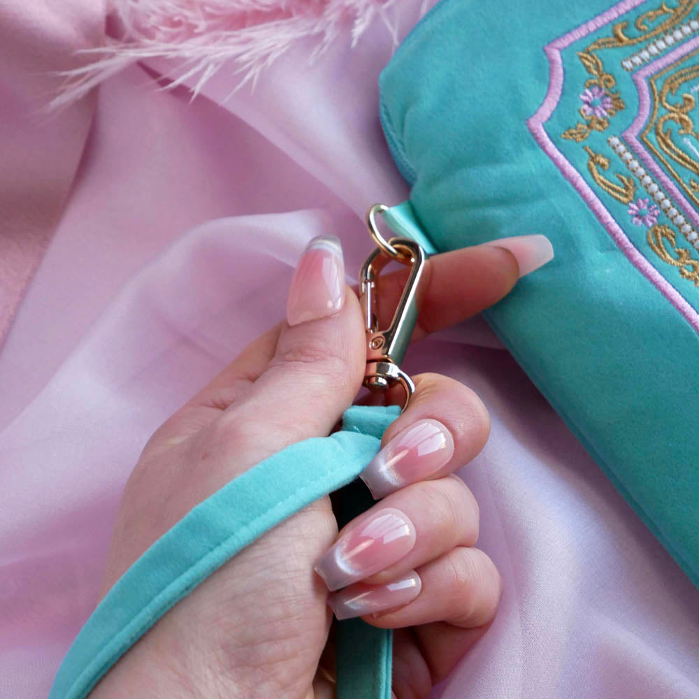 A detail of the aqua Kindle sleeve in Antoinette. The mint green velvet eReader case features pink, cream and gold embroidery with a classic book inspired design. Here a hand shows off the wrist strap with gold detailing.