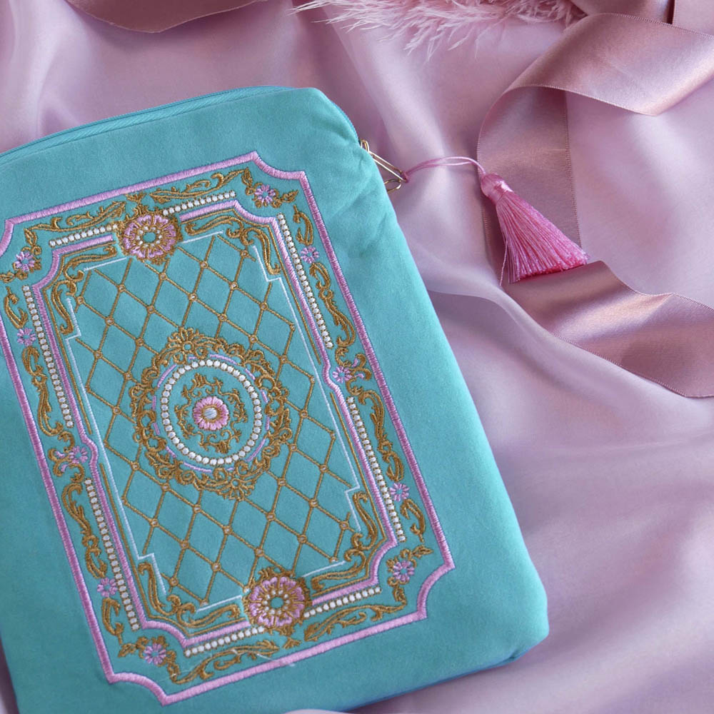 A detail of the aqua Kindle sleeve in Antoinette sits on a pastel pink fabric background. The light green velvet Kobo case features pink, cream and gold embroidery with a classic book inspired design. Here the back of the case shows off the rear pocket and pastel pink tassel.