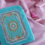A detail of the aqua Kindle sleeve in Antoinette sits on a pastel pink fabric background. The light green velvet Kobo case features pink, cream and gold embroidery with a classic book inspired design. Here the back of the case shows off the rear pocket and pastel pink tassel.