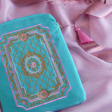 A detail of the aqua Kindle sleeve in Antoinette sits on a pastel pink fabric background. The light green velvet Kobo case features pink, cream and gold embroidery with a classic book inspired design. Here the back of the case shows off the rear pocket and pastel pink tassel.