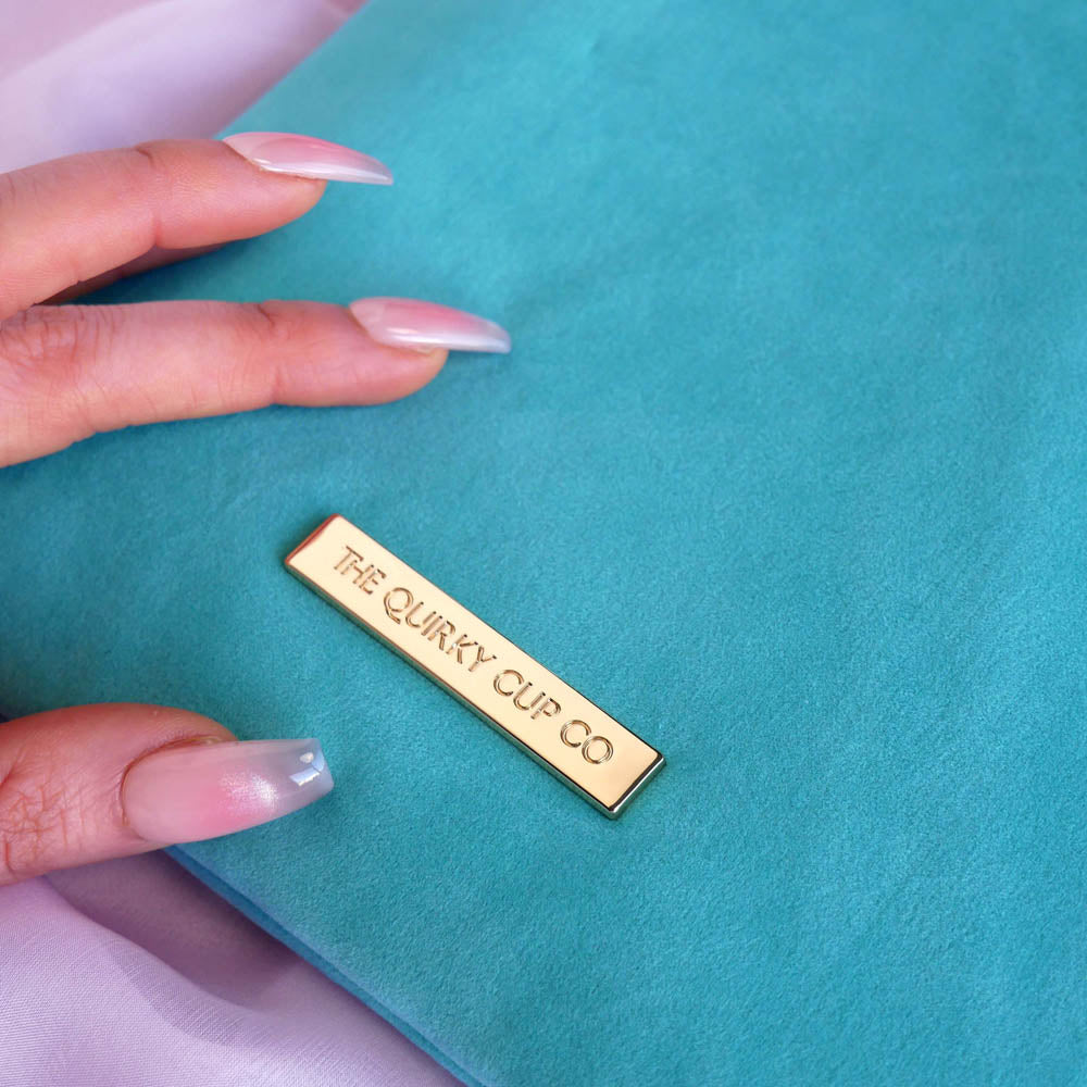 A close-up of the gold metal nameplate engraved with 'THE QUIRKY CUP CO' on the back of the aqua velvet eReader sleeve in Antoinette.