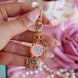 A detail of The gold Antionette Charmlet (A mix and match accessory for decorating your bookish kindle aesthetic) by The Quirky Cup Collective is held by a hand with pastel nails in front of a light pink fabric background. The design featured a gold clasp and multiple charms hanging from different length detachable chains including a floral motif with green and pink enamel details, a bow and a small pink gem.