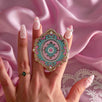 A hand with bejewelled pastel nails is holding a The Quirky Cup Collective Antionette Clingie (it's like a bookish pop socket or enamel phone grip!) in front of a light pink fabric background. This romantasy inspired e-reader product features feminine french inspired design in shades of mint green, pink and white.