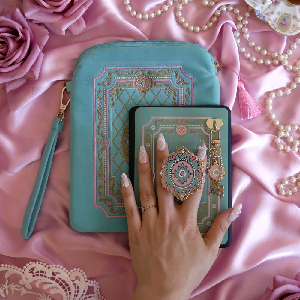 The Antionette E-Reader decor bundle by The Quirky Cup Collective sits on a light pink fabric background, a hand with ornate pastel nails holding it. The set includes an embroidered e-reader case, kindle insert sticker Clingie (imagine an enamel pop socket or decorative phone grip), Charmlet (An accessory with multiple charms attached) and a dust plug. All of the elements feature the same romantic aesthetic with a flower frame aesthetic in shades of mint green, pink, gold and cream.