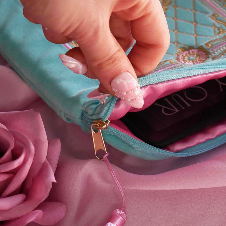 A detail of the Antionette embroidered e-reader case by The Quirky Cup Collective sits on a light pink fabric background, a hand with pastel nails holding it. The feminine kindle case features a wrist strap and pink tassel. The mint green velvet kindle case features pink, gold and cream embroidery of a French-inspired floral frame motif. 