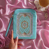 The Antionette embroidered e-reader case by The Quirky Cup Collective sits on a light pink fabric background surrounded by a teacup and pearls , a hand with pastel nails holding it. The kindle case features a wrist strap and pink tassel. The mint green velvet kindle case features pink, gold and cream embroidery of a French-inspired floral frame motif. 