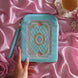 The Antionette embroidered e-reader case by The Quirky Cup Collective sits on a light pink fabric background surrounded by a teacup and pearls , a hand with pastel nails holding it. The kindle case features a wrist strap and pink tassel. The mint green velvet kindle case features pink, gold and cream embroidery of a French-inspired floral frame motif. 
