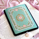 The Antionette Kindle Sticker Insert by The Quirky Cup Collective is attached to a kindle sitting on a book on a light pink fabric background. A sticker to attach directly to your feminine romantasy inspired kindle, the design features a french flower frame motif in shads of gold, pink and cream on the light mint coloured background. The kindle also features a matching Antionette dust plug with the same flower motif.