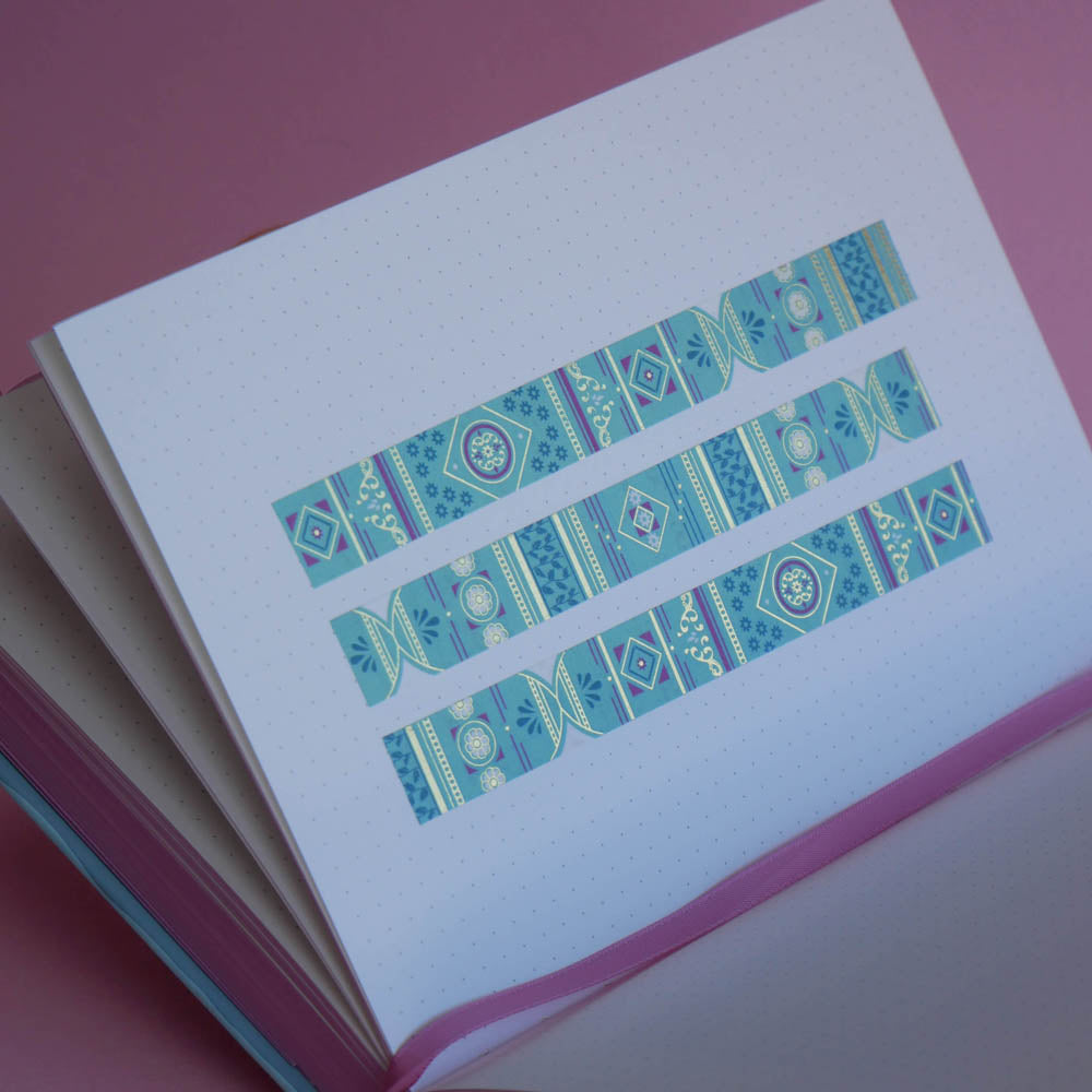 The Aurum washi tape in Antoinette features geometric designs in shades of pink, gold and blue. Three stripes of the tape are in a page of the french inspired notebook against a pink background.