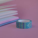 The Aurum design of French inspired washi tape as part of the Antoinette collection sits on a light pink background. The whimsical washi tape features geometric flower motifs in shades of blue, pink and foil gold.