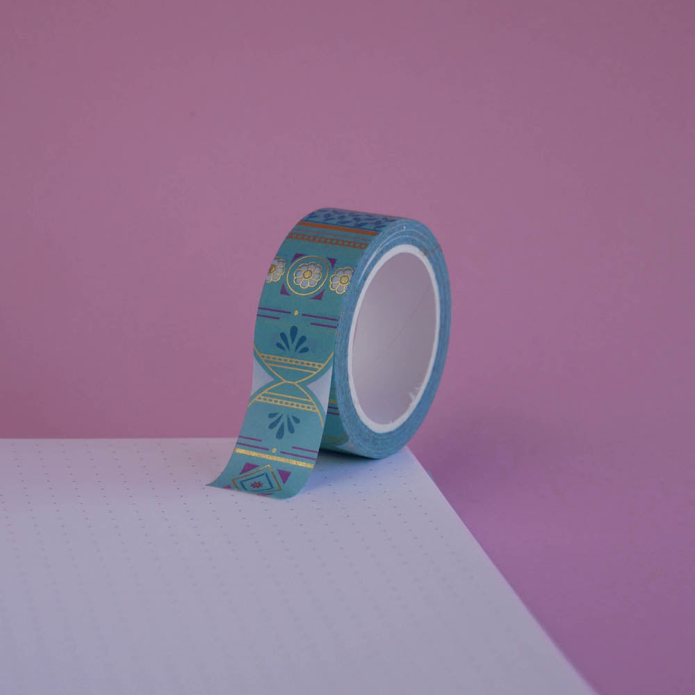 The Aurum design of French inspired washi tape as part of the Antoinette collection sits on a light pink background. The whimsical washi tape features geometric flower motifs in shades of blue, pink and foil gold.