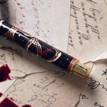 The Quirky Cup Collective Bad Blood pen features an intricate snake motif in red, gold and black. The pen sits on a background of handwritten calligraphy notes and spilt red wax.