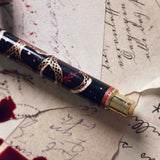 The Quirky Cup Collective Bad Blood pen features an intricate snake motif in red, gold and black. The pen sits on a background of handwritten calligraphy notes and spilt red wax.