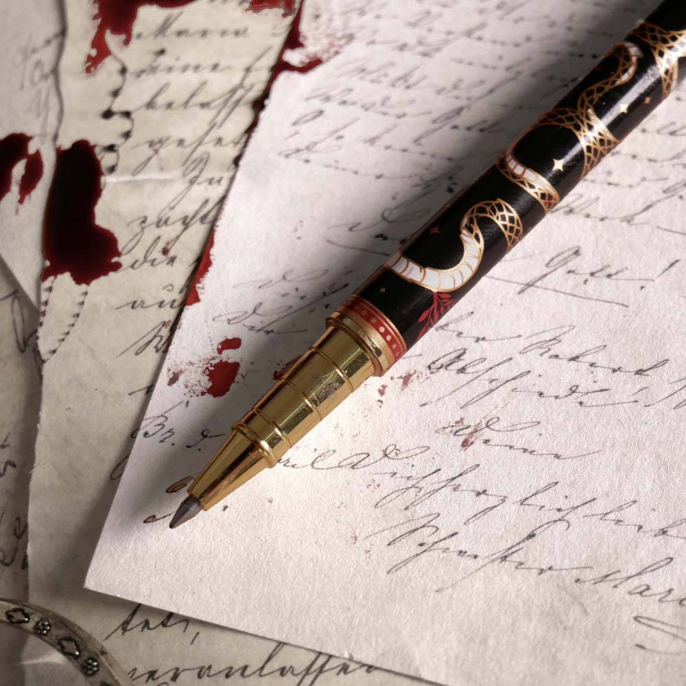 The Quirky Cup Collective Bad Blood pen features an intricate snake motif in red, gold and black and an engraved 'Bad Blood' text on the clip. The pen sits on a background of handwritten calligraphy notes and spilt red wax.