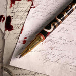 The Quirky Cup Collective Bad Blood pen features an intricate snake motif in red, gold and black and an engraved 'Bad Blood' text on the clip. The pen sits on a background of handwritten calligraphy notes and spilt red wax.