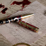 The Quirky Cup Collective Bad Blood pen features an intricate snake motif in red, gold and black and an engraved 'Bad Blood' text on the clip. The pen sits on a background of handwritten calligraphy notes and spilt red wax.