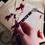 The Quirky Cup Collective Bad Blood pen features an intricate snake motif in red, gold and black and an engraved 'Bad Blood' text on the clip. A hand with long, intricate red nails holds the pen on a background of handwritten calligraphy notes and spilt red wax.