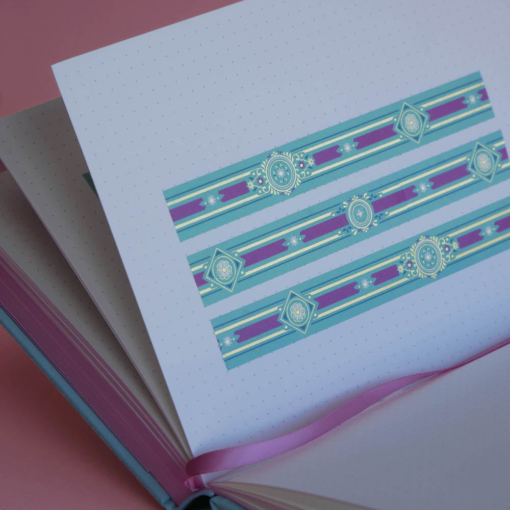 The Balteus washi tape in Antoinette features classic book inspired designs in shades of pink, hold and blue. Three stripes of the tape are in a page of the french inspired notebook against a pink background.