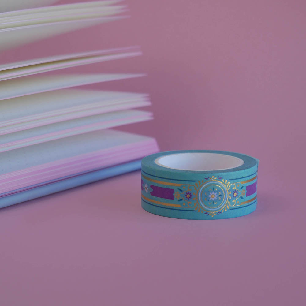 The Balteus design of French inspired washi tape as part of the Antoinette collection sits on a light pink background. The romantasy inspired washi tape features a classical book inspired motif in shades of blue, pink and foil gold.